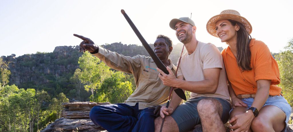 Indigenous guide points across a rugged escarpment while sharing cultural knowledge with two smiling visitors, one holding a traditional spear, in a bushland setting.