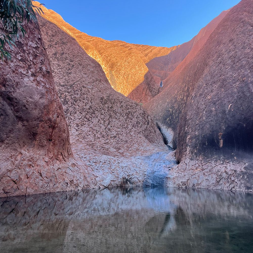 Narrow waterhole between towering red sandstone walls, reflecting the rugged rock formations beneath a clear blue sky.