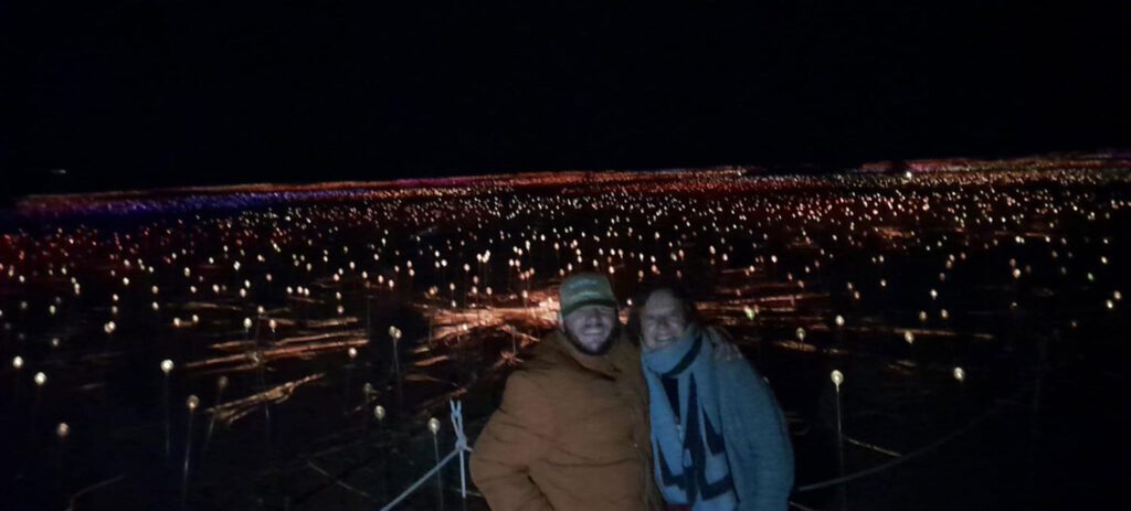 Two visitors pose together at night amid a vast illuminated field of colorful lights stretching across the desert landscape.