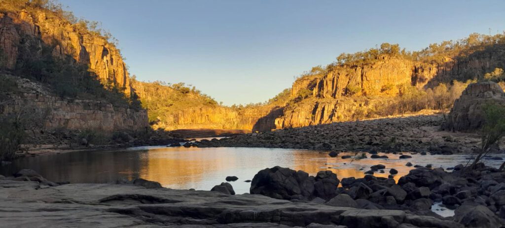 Calm waterhole surrounded by rugged rocky gorge walls glowing in warm sunlight, with scattered boulders and a clear blue sky