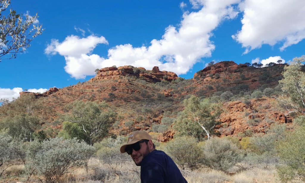 Smiling visitor wearing sunglasses and a cap poses in arid bushland beneath rugged red rock hills and a vivid blue sky with white clouds.
