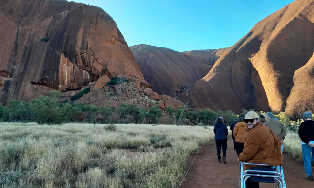 Visitor using a mobility aid walks along a red dirt trail at the base of Uluru, surrounded by native grassland and towering sandstone formations.