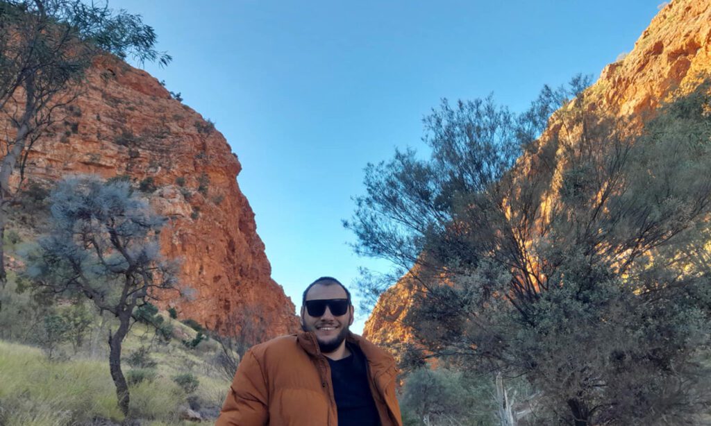 Smiling visitor wearing sunglasses and an orange jacket stands on a bushland trail between towering red rock cliffs beneath a clear blue sky.