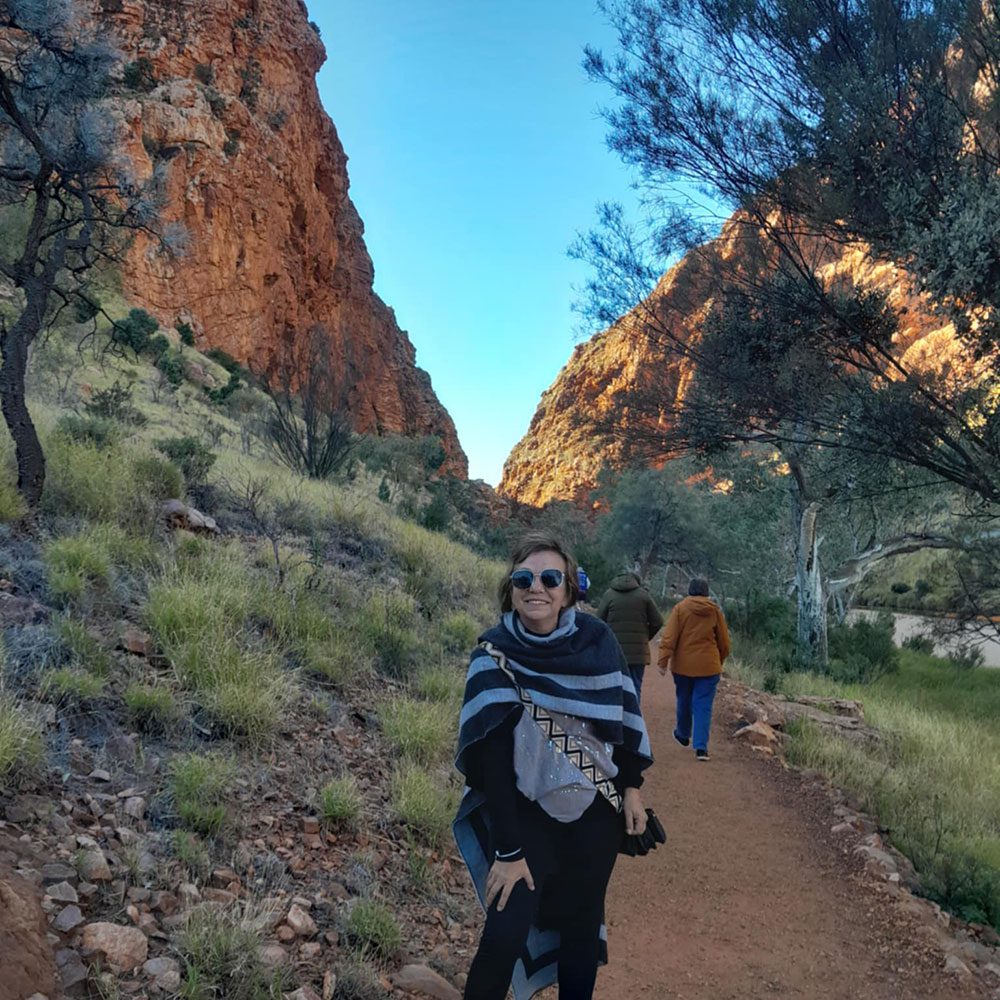 Smiling visitor wearing sunglasses and a striped shawl stands on a red dirt trail between rugged red rock cliffs and native bushland