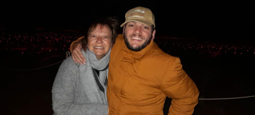 Smiling man in an orange jacket and cap embraces a woman in a gray coat at night, with a field of red lights glowing in the background.