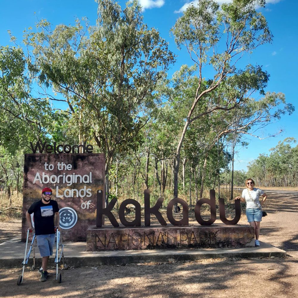 Two visitors pose beside a “Welcome to the Aboriginal Lands of Kakadu” sign in a eucalyptus woodland under a bright blue sky.