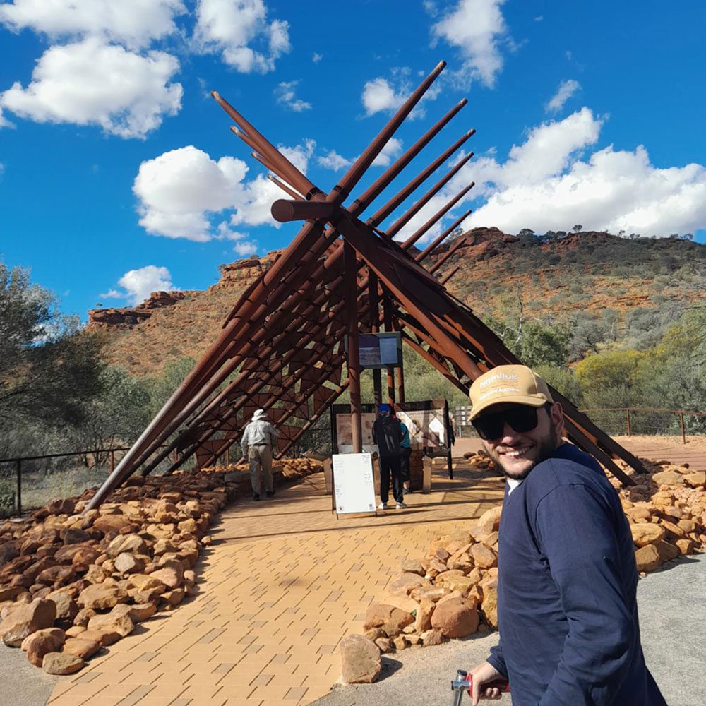 Visitor using a mobility aid poses near a large angular timber entrance structure, with rugged red cliffs and a bright blue sky in the background.