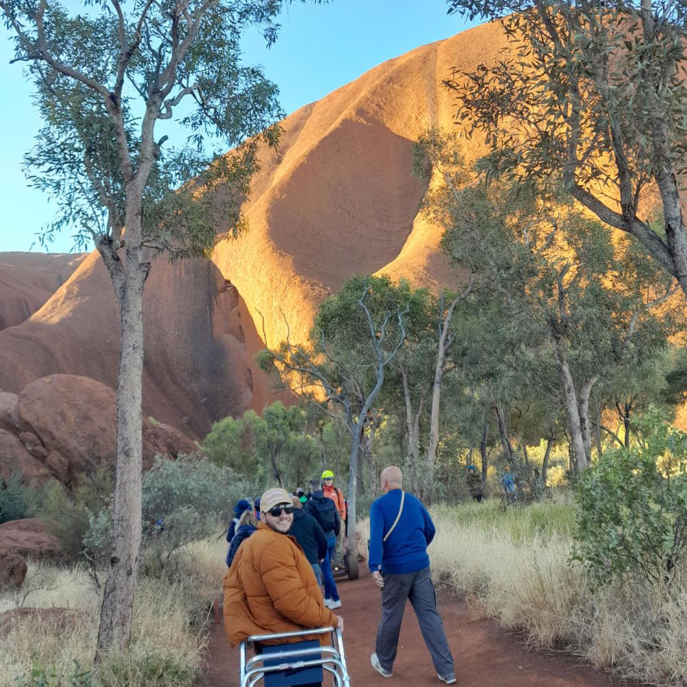 : Visitors walk along a red dirt trail through native bushland at the base of Uluru, with one person using a mobility aid.
