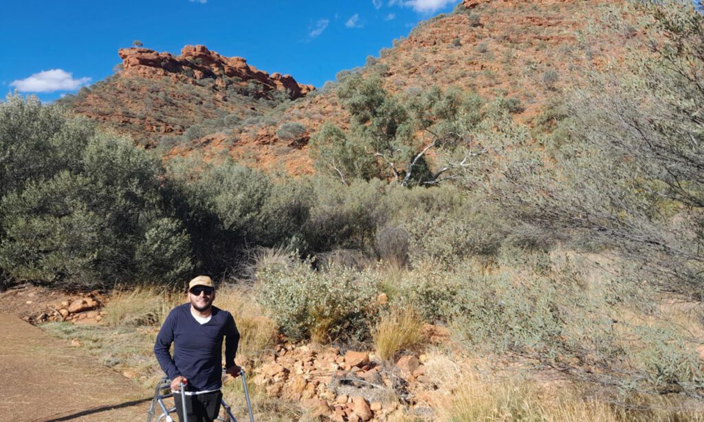 Visitor using a mobility aid walks along a bush trail surrounded by native vegetation and rugged red rock hills beneath a bright blue sky.