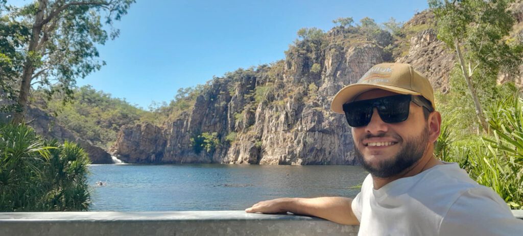 Smiling visitor wearing sunglasses and a cap poses beside a clear blue gorge pool surrounded by rugged cliffs and native vegetation.