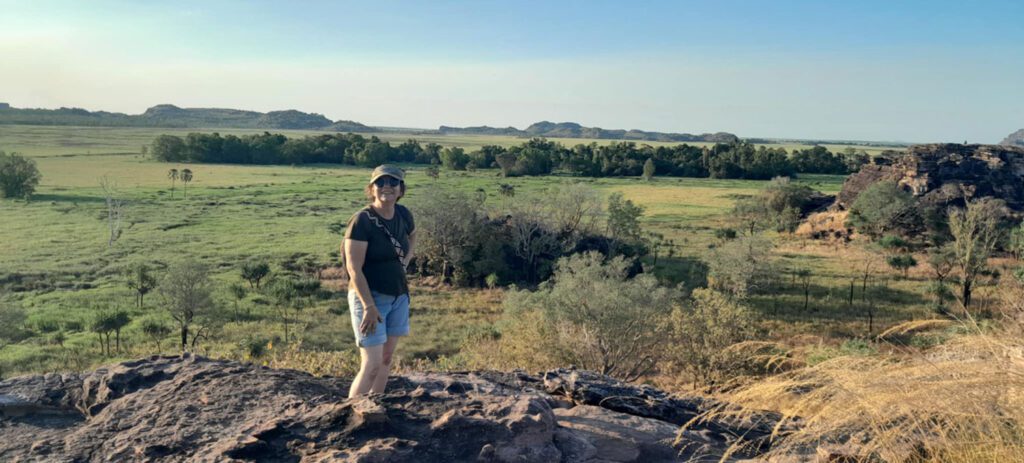 Visitor stands on a rocky lookout overlooking expansive green wetlands, scattered trees, and distant sandstone formations beneath a clear blue sky.