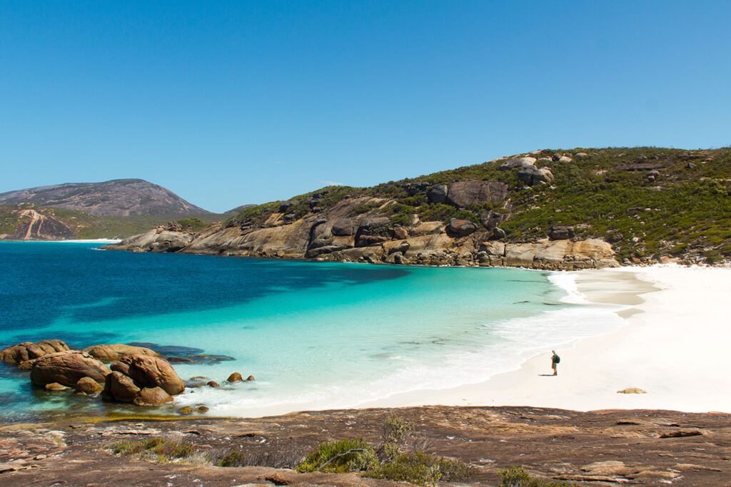 A lone person standing on pure white sand looking out over a narrow inlet to the ocean over crystal clear vibrant turquoise water with rocky hills in the background in Western Australia