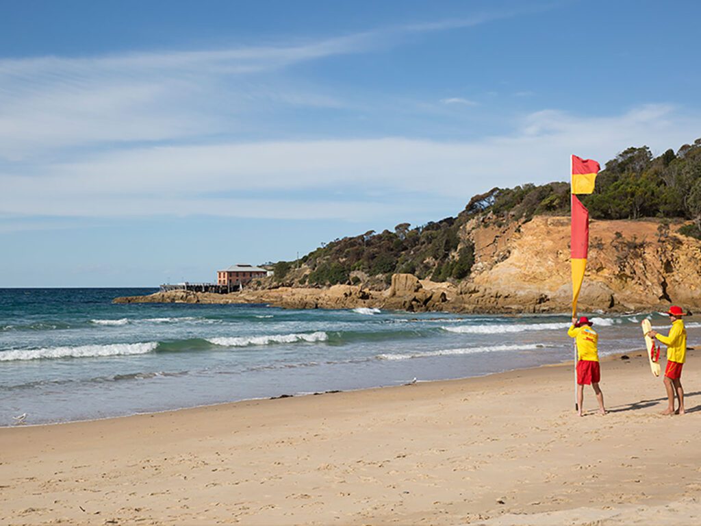 Two lifeguards erecting a red and yellow flag on a sand beach with small waves and a hill with trees in the background