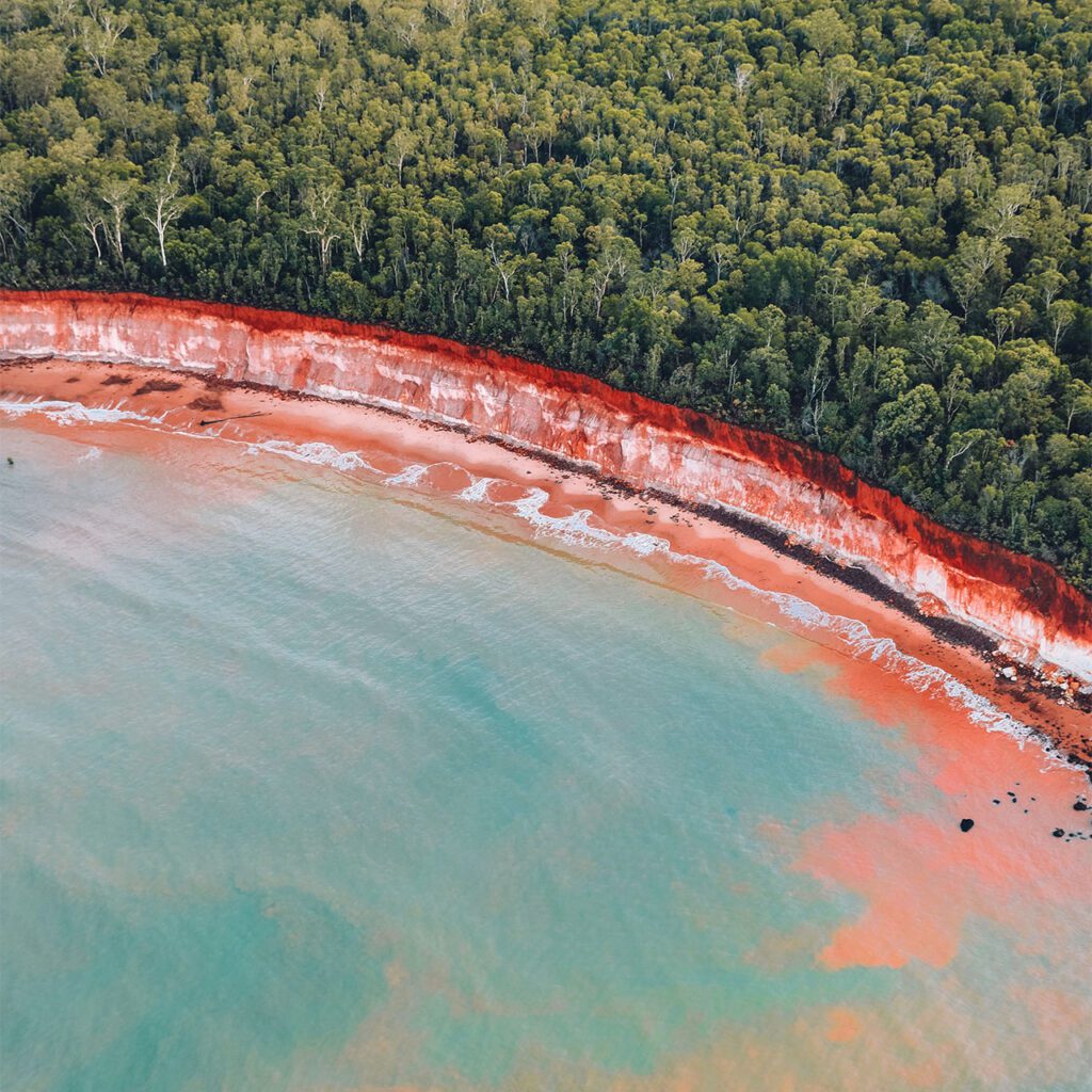 Candy blue coloured water lapping against a narrow pink stretch of sand which is topped by a sheer cliff of brilliant vermillion rocks with native trees which reach the very edge of the cliff in South Australia