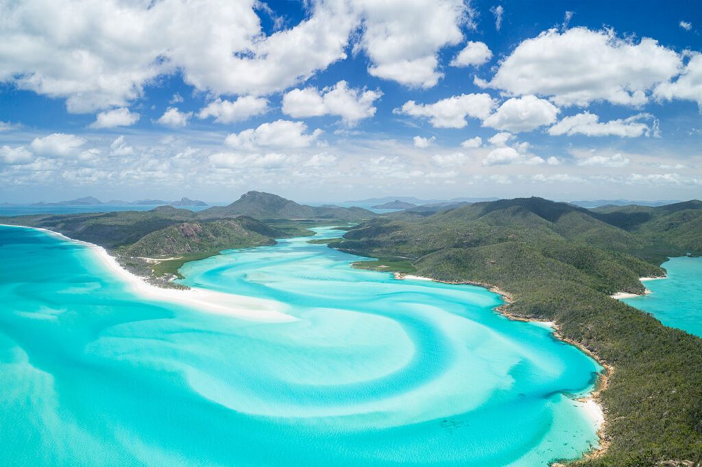 A pale blue waterway winding through pure white sand, framed by lush green mountains in the background, capturing the breathtaking beauty of the Whitsunday Islands in Queensland, Australia
