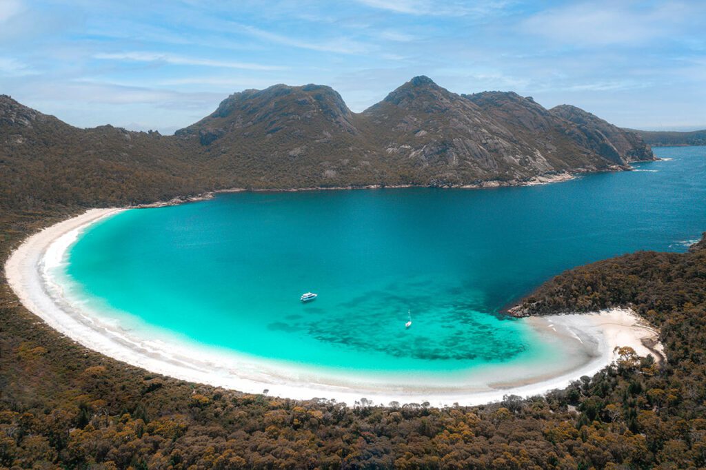 A horseshoe shaped bay with a small boat anchored on the turquoise water with rugged tree-covered mountains in the background in Wineglass Bay Tasmania